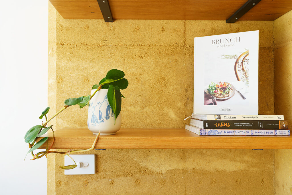 Wooden bookshelf in a rammed earth home with a growing indoor plant and books on sustainability and cooking